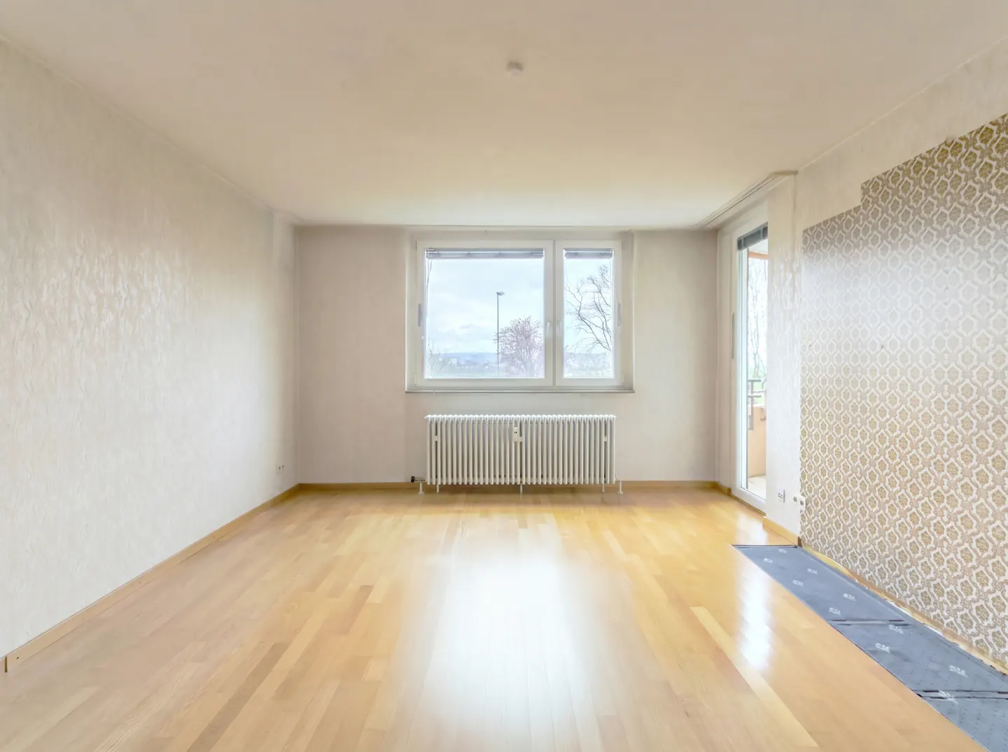 Bright, empty room with light wood floors, white walls, and a window. A radiator sits beneath the window. One wall has patterned wallpaper.
