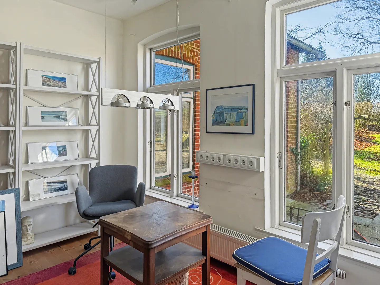 Bright room with a gray chair, wooden table, and white bookshelf with framed art. Large windows offer views of the outdoors.