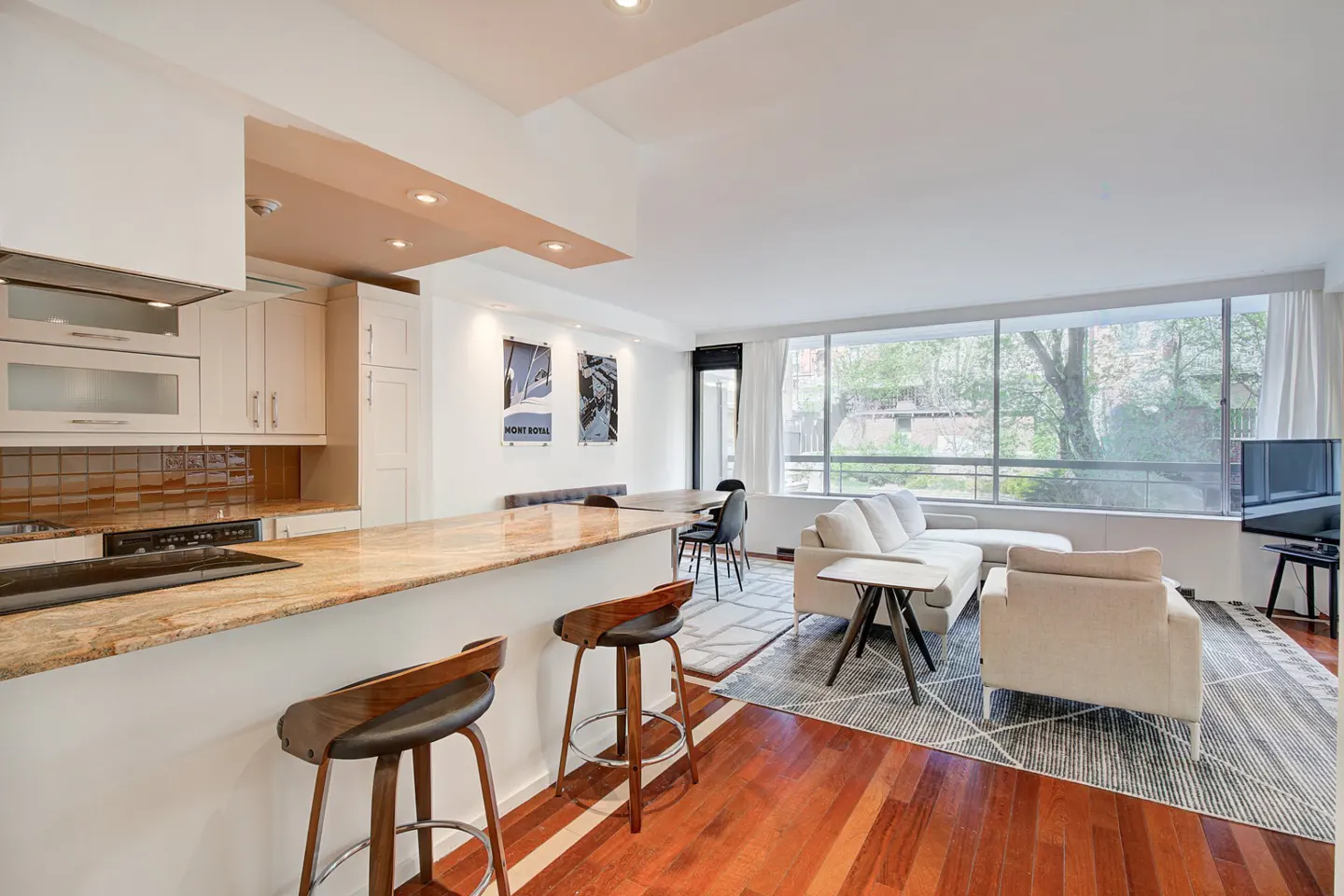 Open-concept living space with kitchen bar, stools, and seating area with large windows. Hardwood floors and neutral color palette.