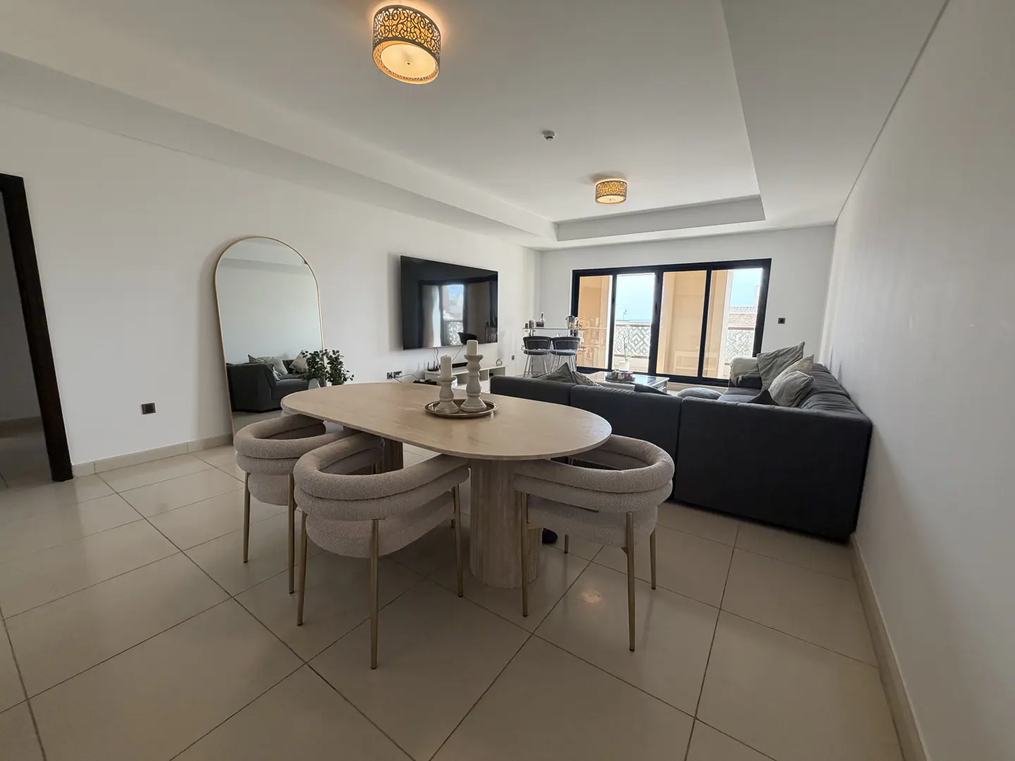 Bright, modern living room with a light wood dining table, four chairs, and a gray sectional sofa. A large mirror reflects the room.