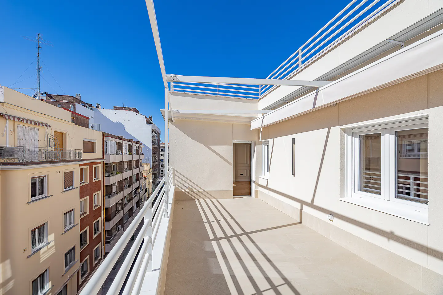 A bright, sunny balcony with white railings overlooks city buildings under a clear blue sky.