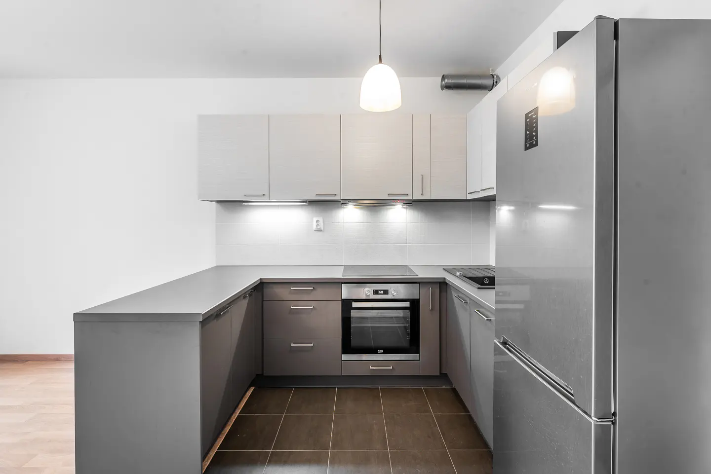 Modern U-shaped kitchen with grey cabinets, stainless steel oven, and a tall silver refrigerator on dark tiled floors.