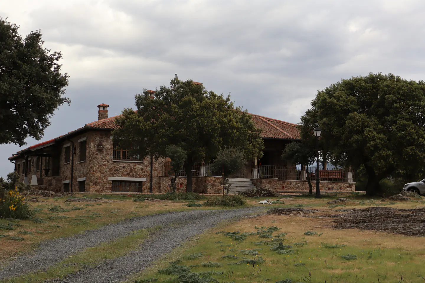 Stone house with a red tile roof and chimney, surrounded by trees and a gravel driveway under a cloudy sky.