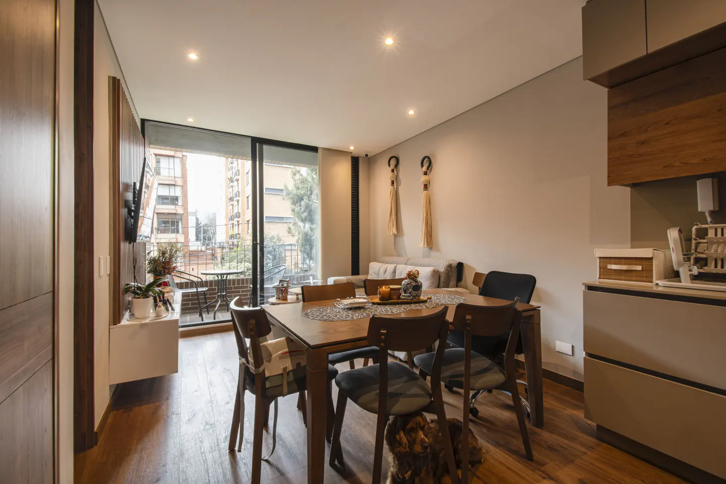 A modern apartment interior with a wooden dining table and chairs, a balcony with a city view, and a kitchen area.