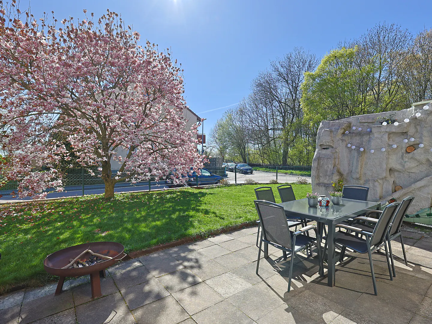 Outdoor patio with a gray table and chairs, a fire pit, and a blooming pink tree on a sunny day.