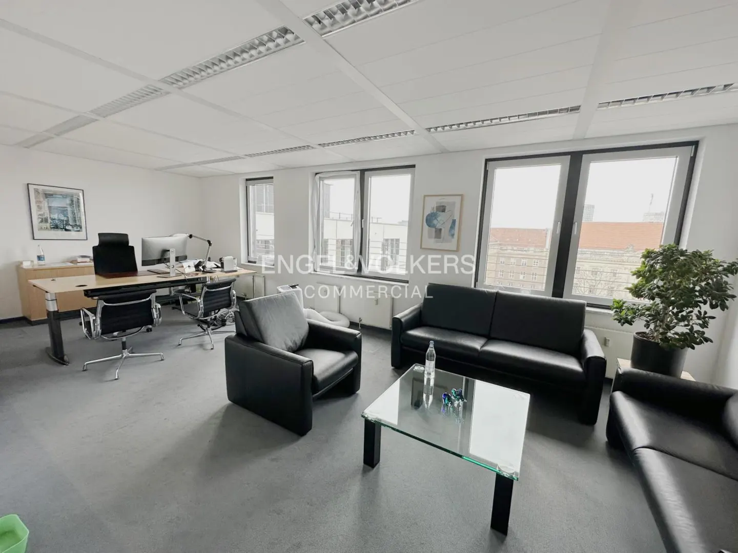 Bright office space with black leather seating, glass table, and desk with computer. Windows show city view. "Engel & Volkers Commercial" logo visible.