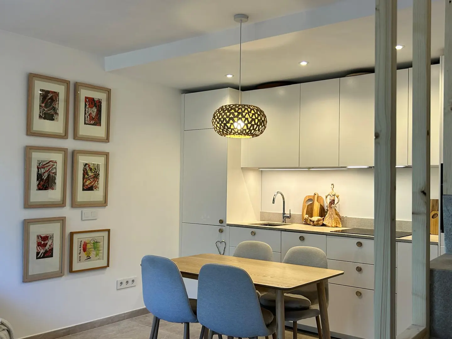 A modern kitchen with white cabinets, a wooden table with blue chairs, and framed art on the wall. A decorative pendant light hangs above the table.