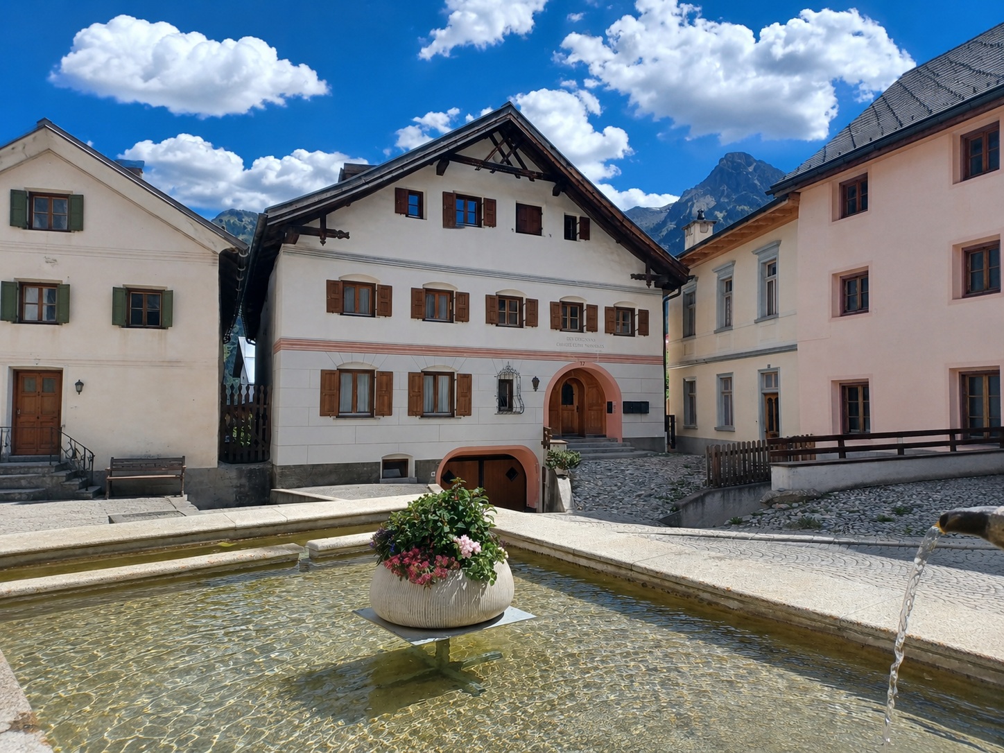A stone fountain with a flower planter sits before three traditional European buildings under a bright, cloudy blue sky.