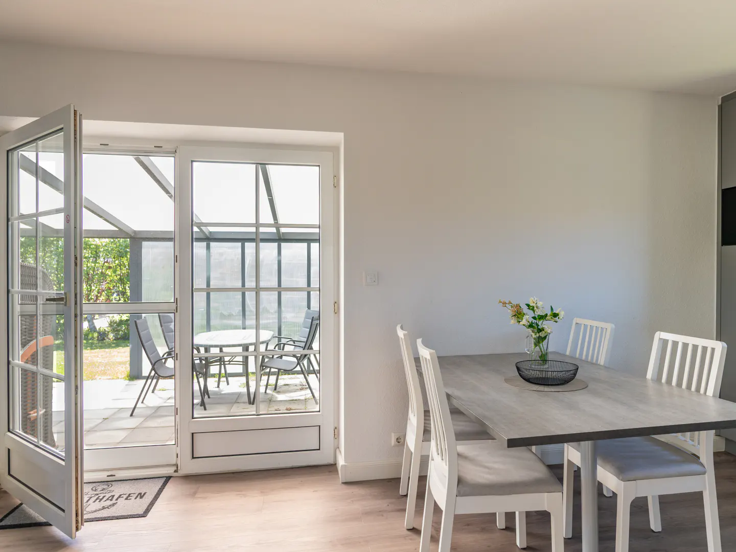 Dining area with a light wood table, white chairs, and a vase, opening to a sunroom with patio furniture.