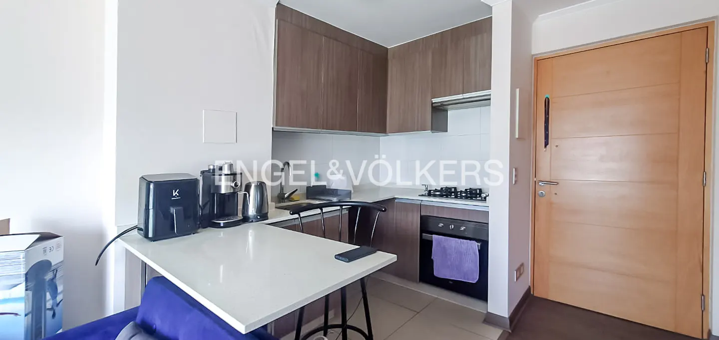 A modern kitchen with brown cabinets, white countertops, and stainless steel appliances. A white table with a black chair is in the foreground. A light wood door is on the right.
