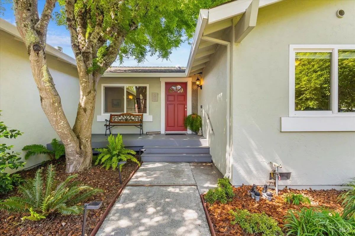 Exterior view of a single-story house with a red front door, gray siding, and a concrete walkway lined with mulch and plants.