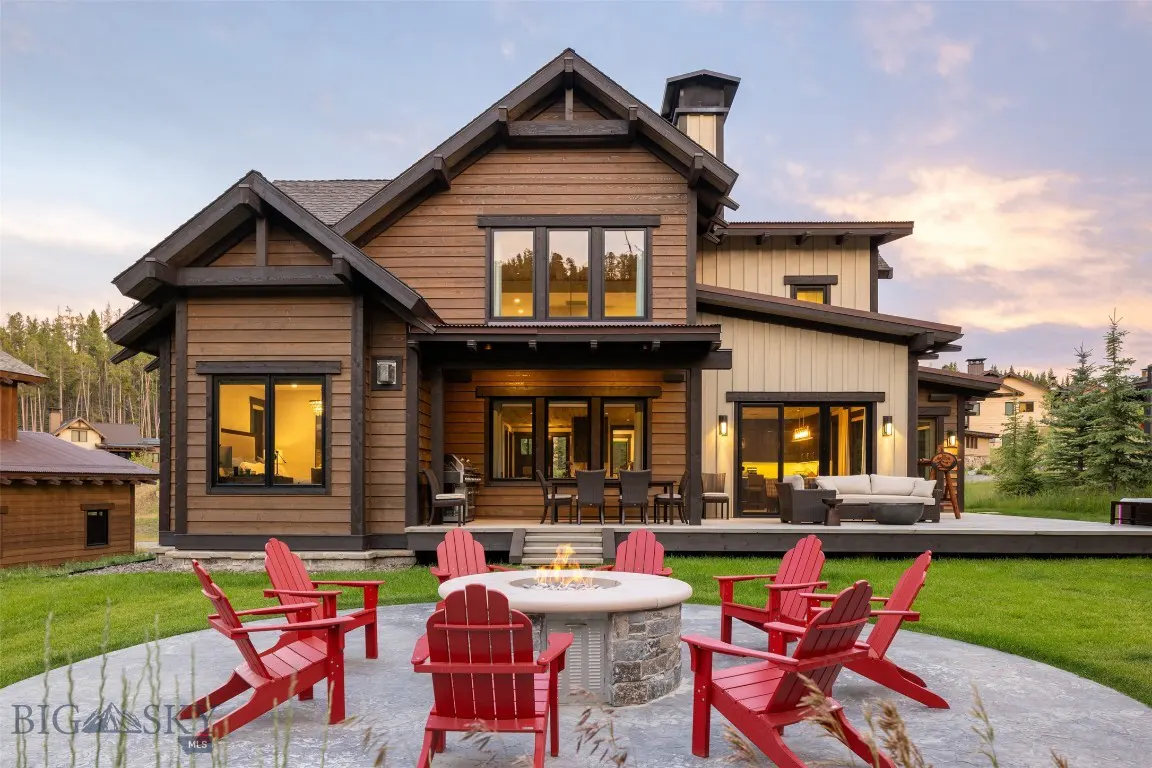 Two-story brown house with a stone fire pit and red Adirondack chairs on a concrete patio. A deck with outdoor furniture is visible.