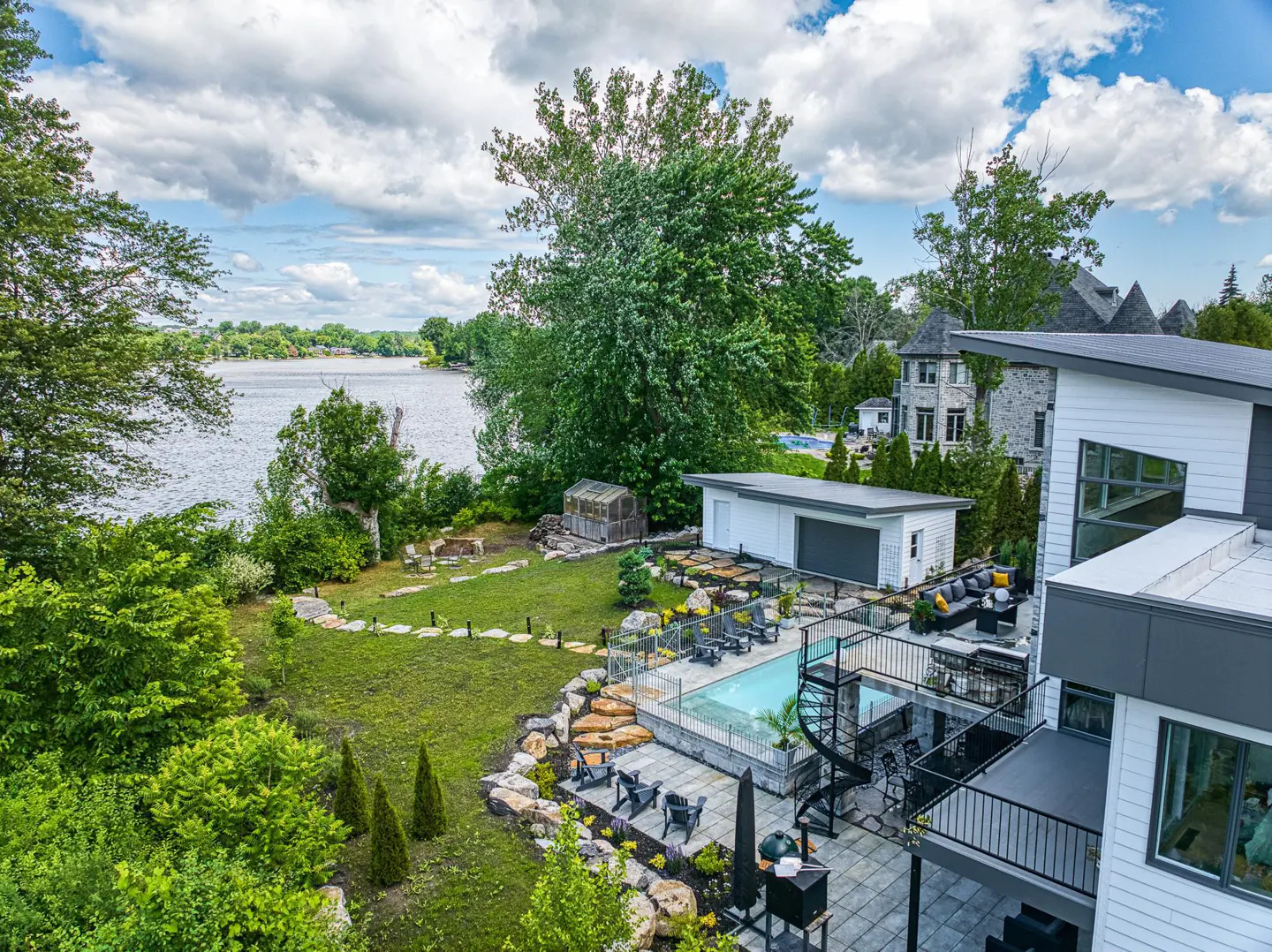 Aerial view of a modern home with a pool, patio, and lake access. Lush green lawn, trees, and blue sky with clouds.