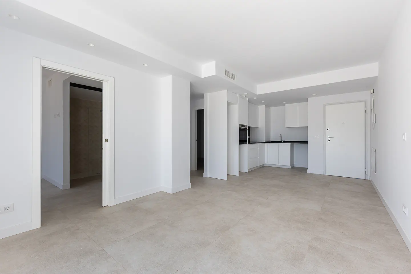 Minimalist open-plan apartment with white walls, light tile floors, a modern white kitchen, and a closed entry door.