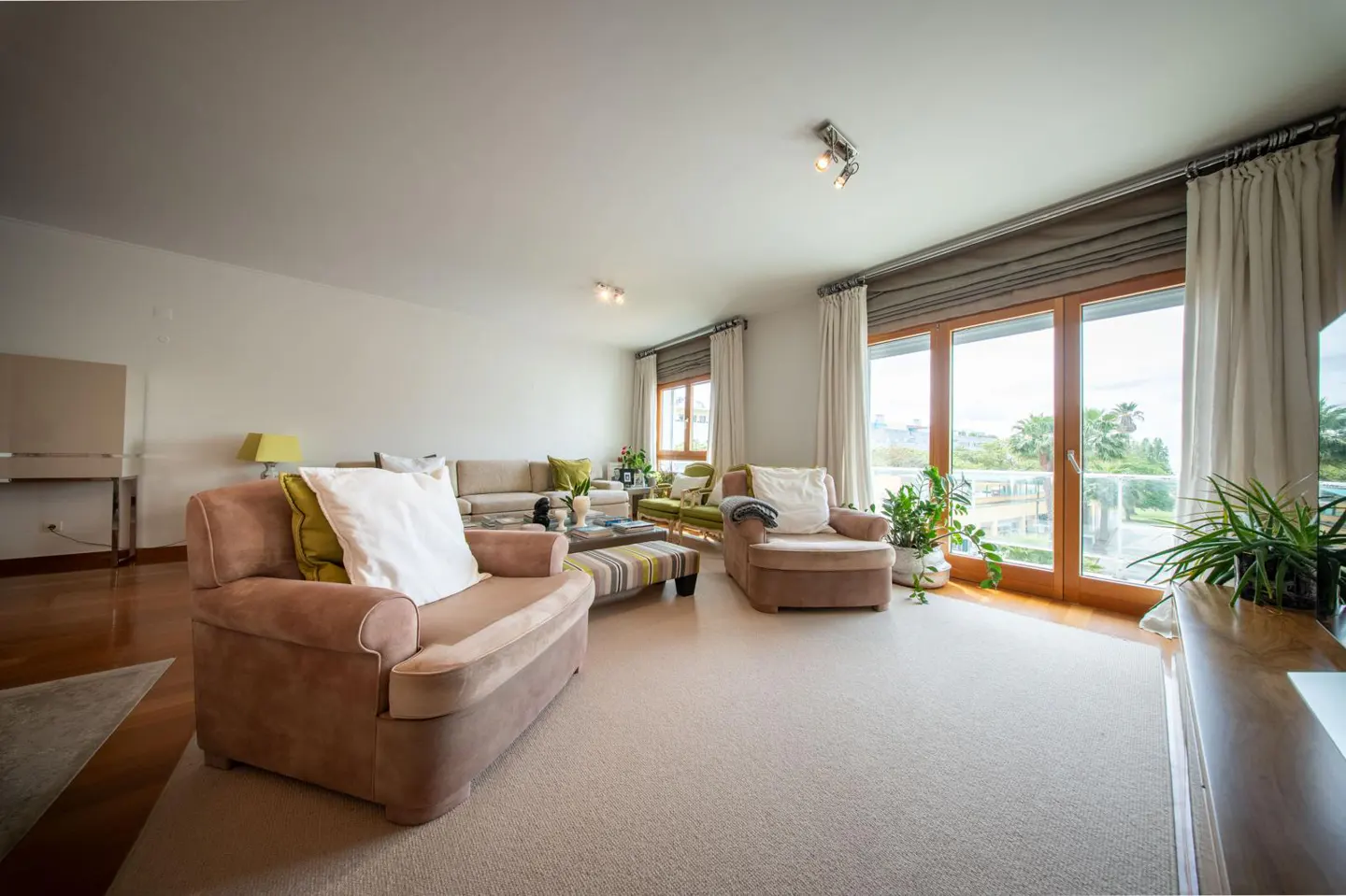 Bright living room with beige carpet, sofa, armchairs, and large windows with curtains. Plants add a touch of nature.