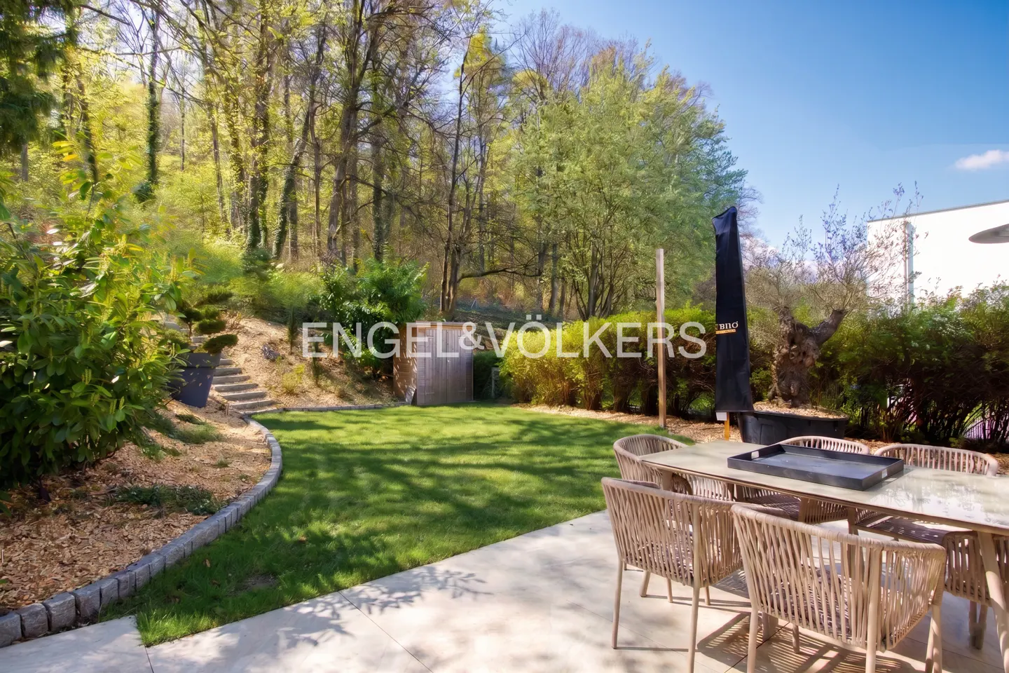 Outdoor patio with a table and chairs, a green lawn, and a wooded area in the background. The sky is blue.