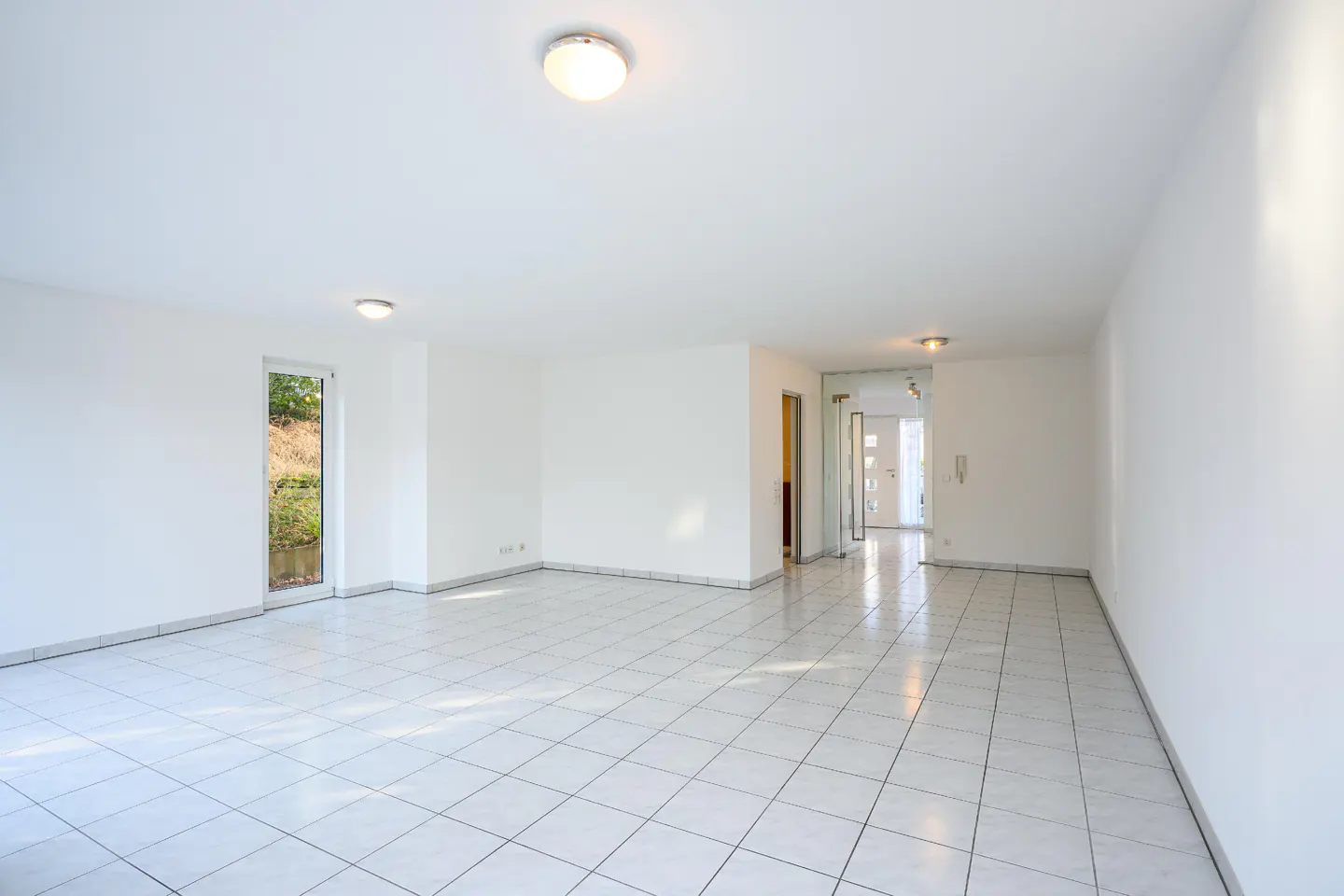 Bright, empty room with white walls and tiled floor. A window shows greenery outside. Hallway visible in the background.