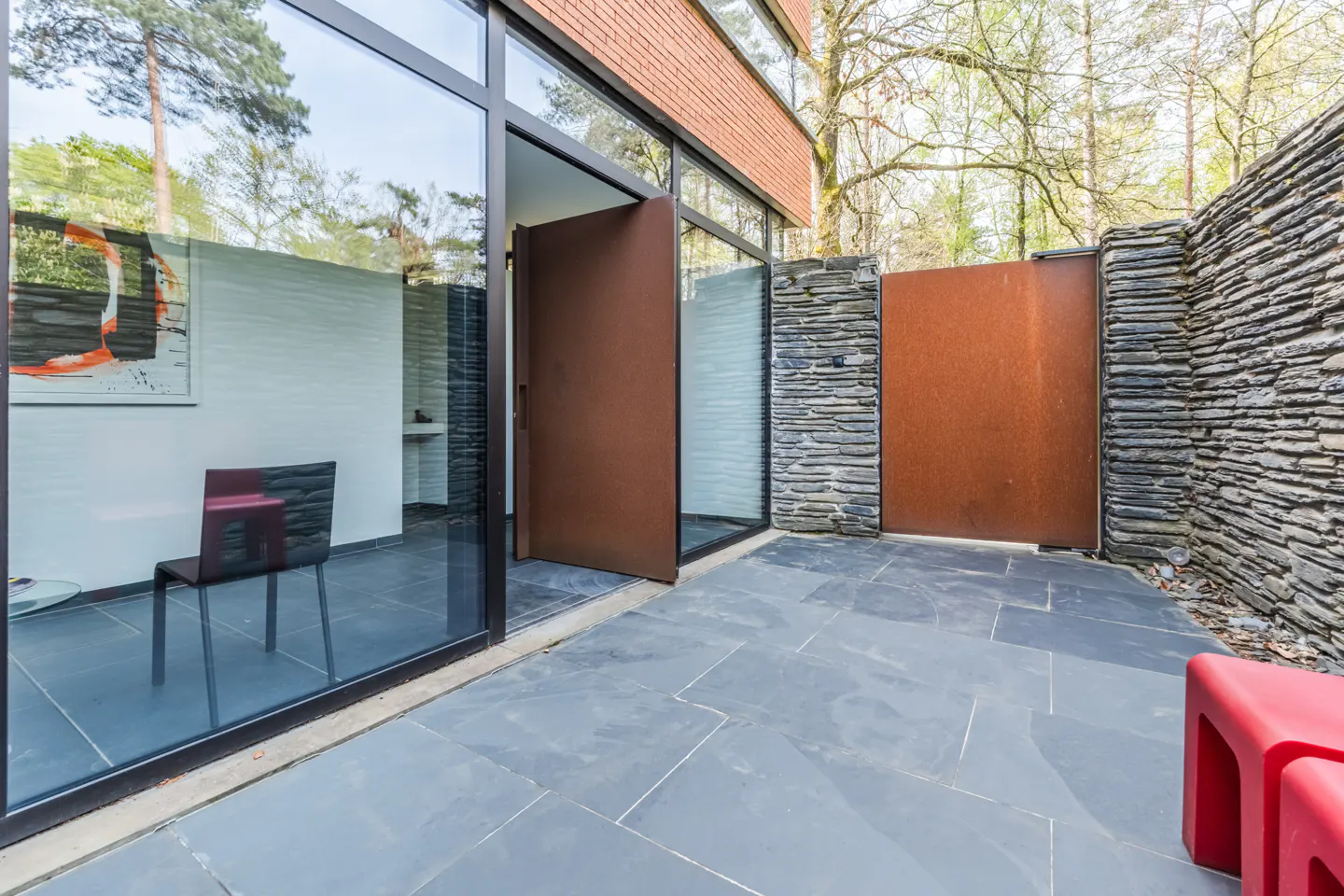 Modern home exterior with a large, open brown door, stone walls, and gray tiled patio. A red chair is visible through the glass wall.