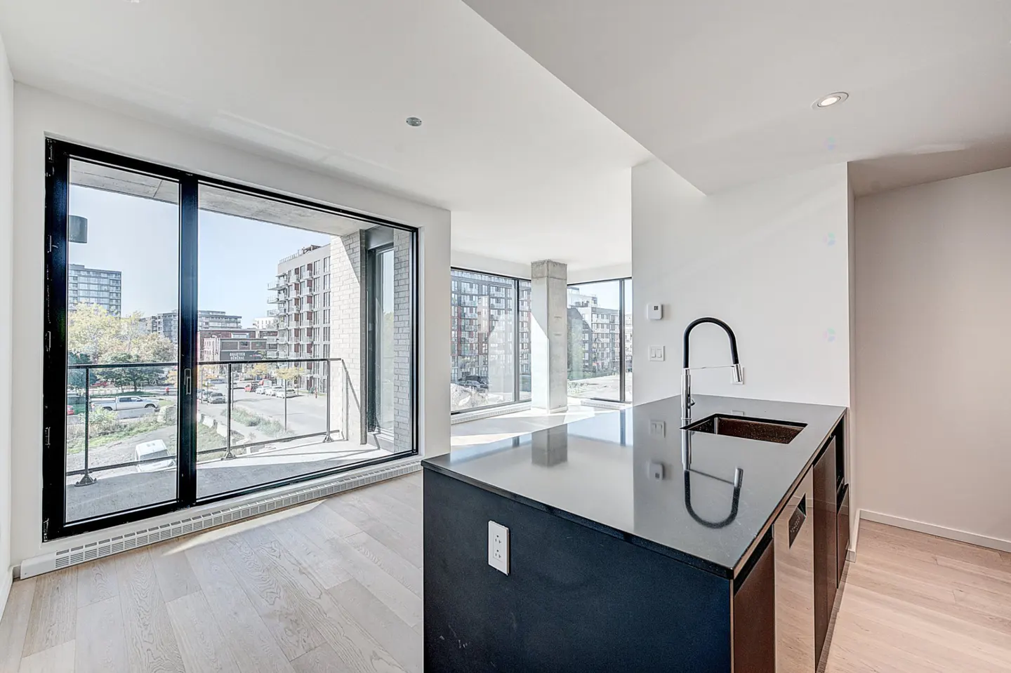 Bright, modern apartment interior with a black kitchen island, sink, and faucet. Large windows offer a city view. Light wood floors and white walls create a clean aesthetic.