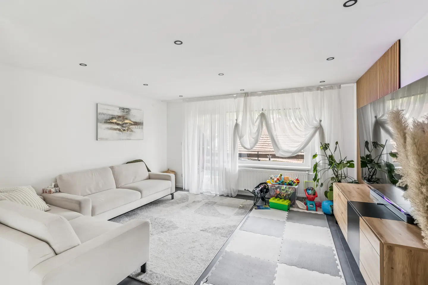 Bright living room with a white sofa, grey patterned rug, sheer curtains, wooden media unit, and a children's play area.