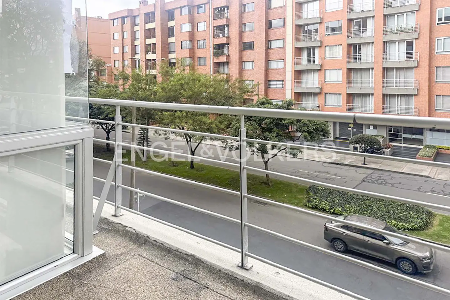 Balcony view of a street with a car, trees, and a brick apartment building in the background.