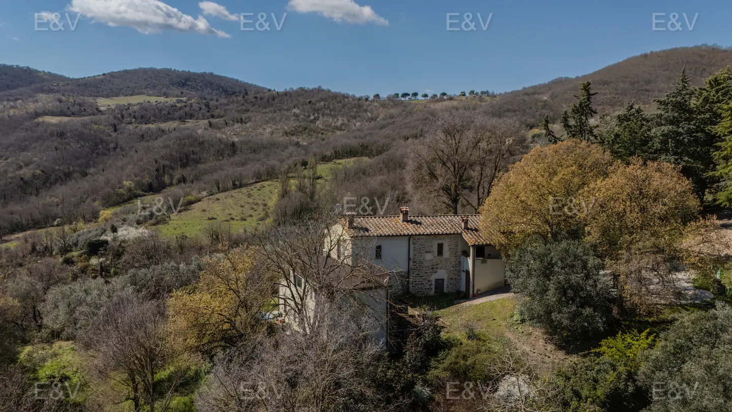 Stone house with a red tile roof nestled in a green, hilly landscape under a blue sky.