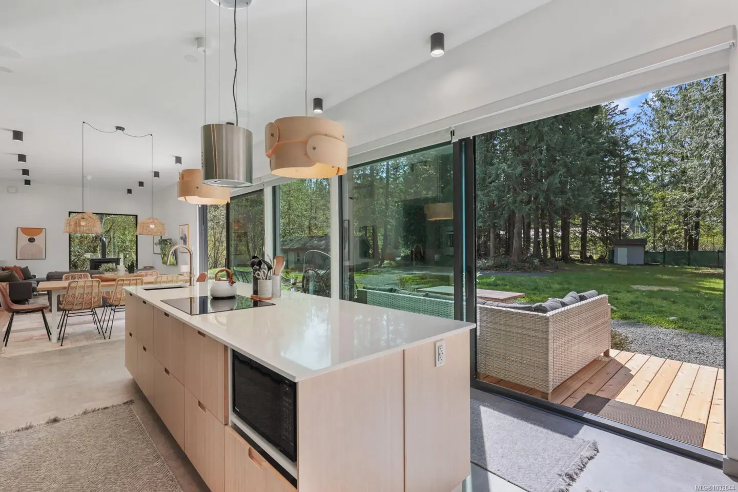 Bright, modern kitchen with a large island, white countertops, and wood cabinets. Sliding glass doors open to a backyard with trees and outdoor seating.
