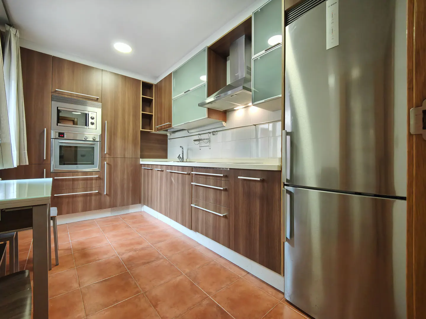 A kitchen with brown cabinets, stainless steel appliances, and terracotta tile flooring.
