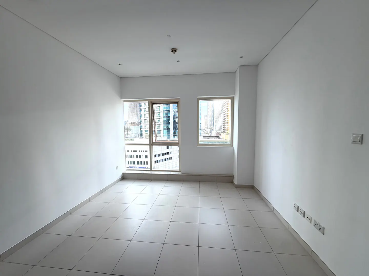 Empty room with white walls, tiled floor, and two windows showing a city view.