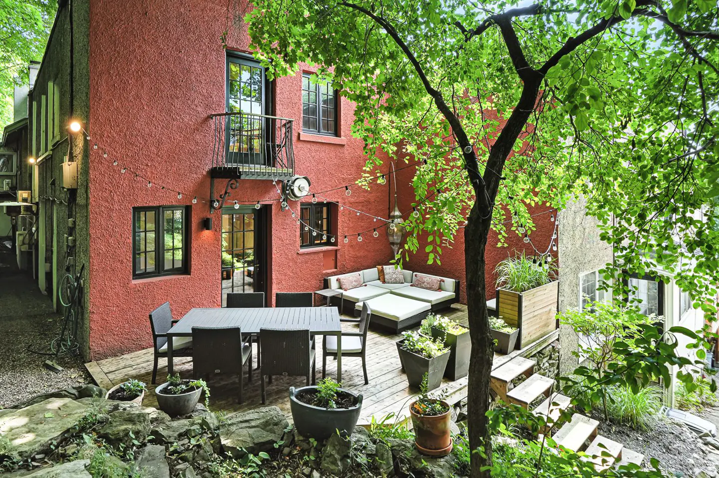 Outdoor patio with a red stucco building backdrop. A dining table, lounge seating, and greenery create a cozy space. String lights add ambiance.