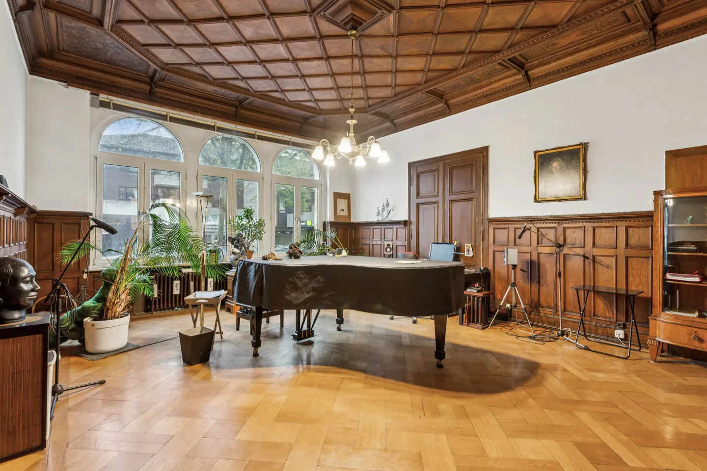 A spacious room with a grand piano, wood paneled walls, herringbone floors, and a coffered wood ceiling.