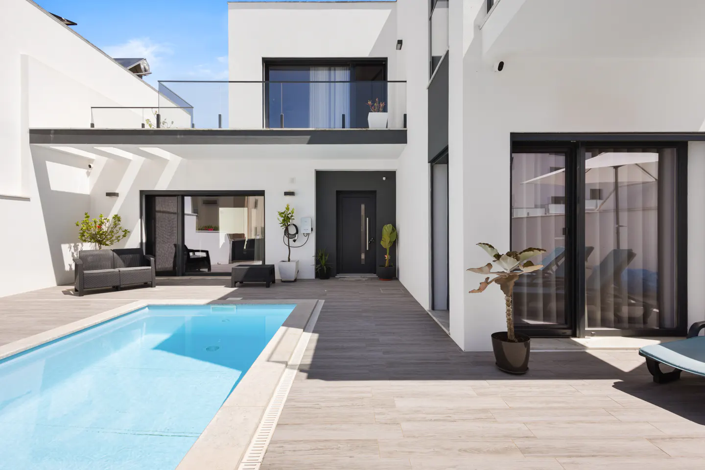 Modern white house with a blue pool and gray patio. Black door and furniture accent the bright exterior. Sunny sky above.