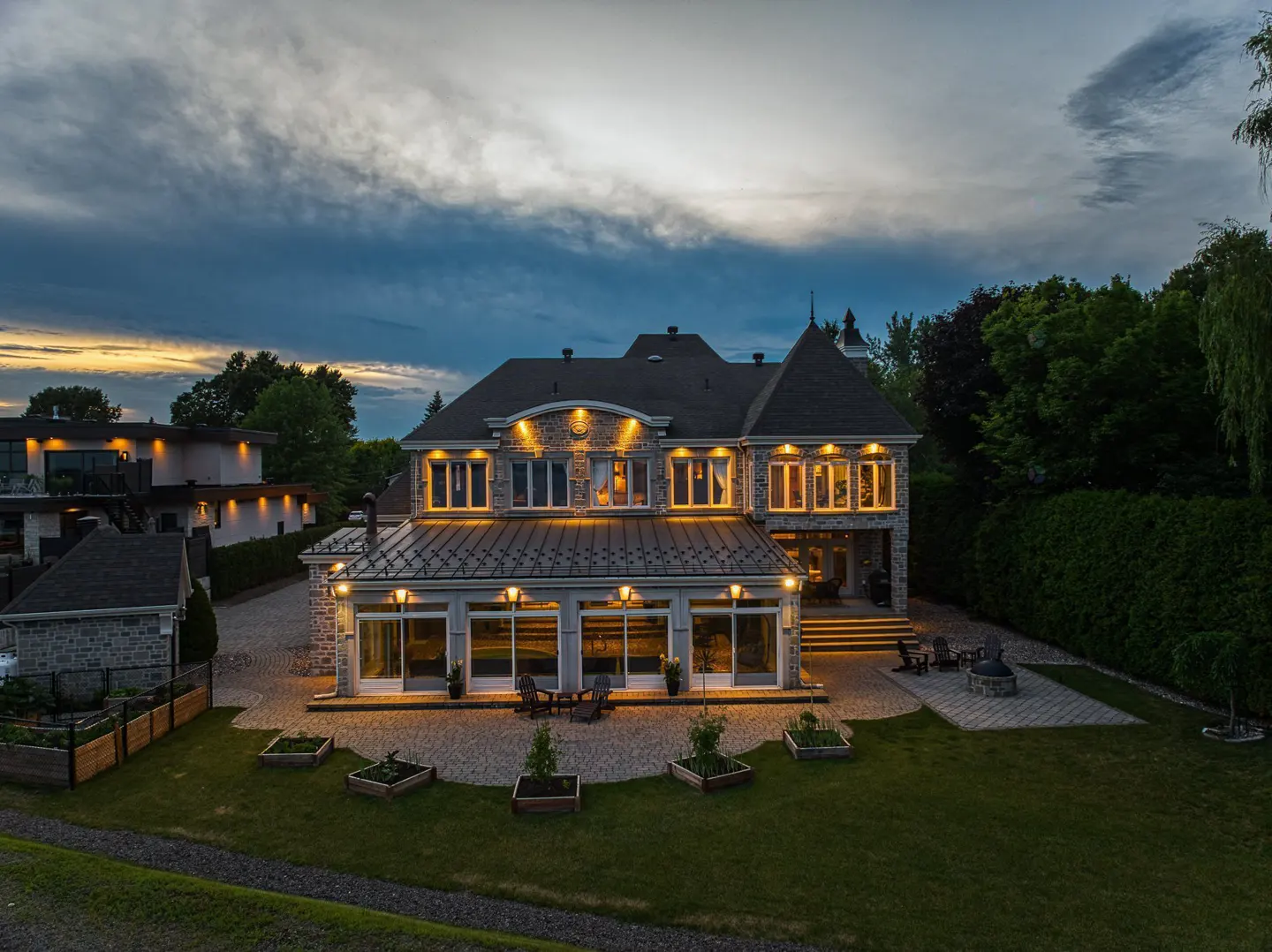 Two-story stone house with a dark roof, illuminated windows, and a green lawn at dusk.