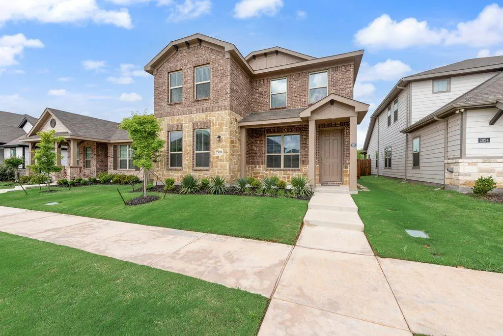 Two-story brick and stone house with a brown front door, green lawn, and concrete walkway under a blue sky.