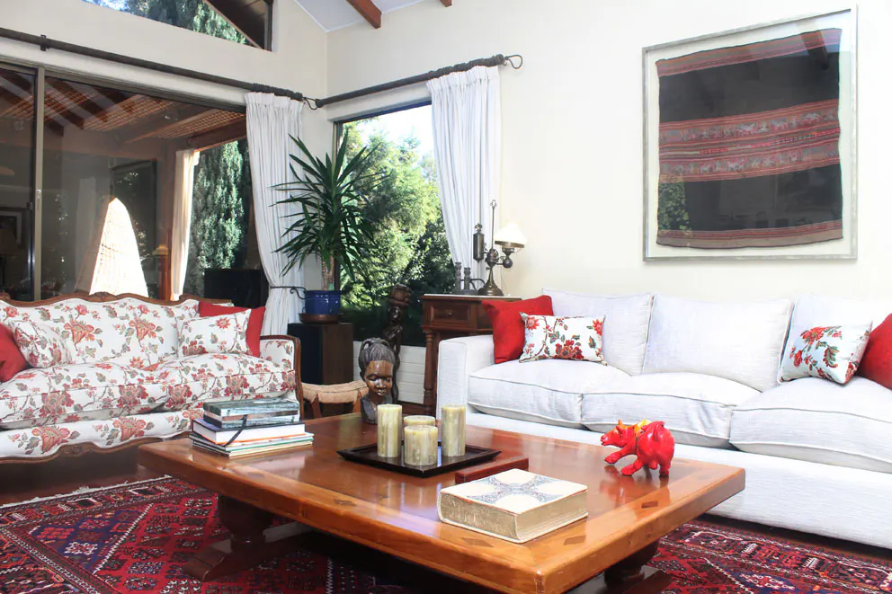 Bright living room with white sofas, floral accents, and a wooden coffee table on a red patterned rug. Large windows offer a view of lush greenery.