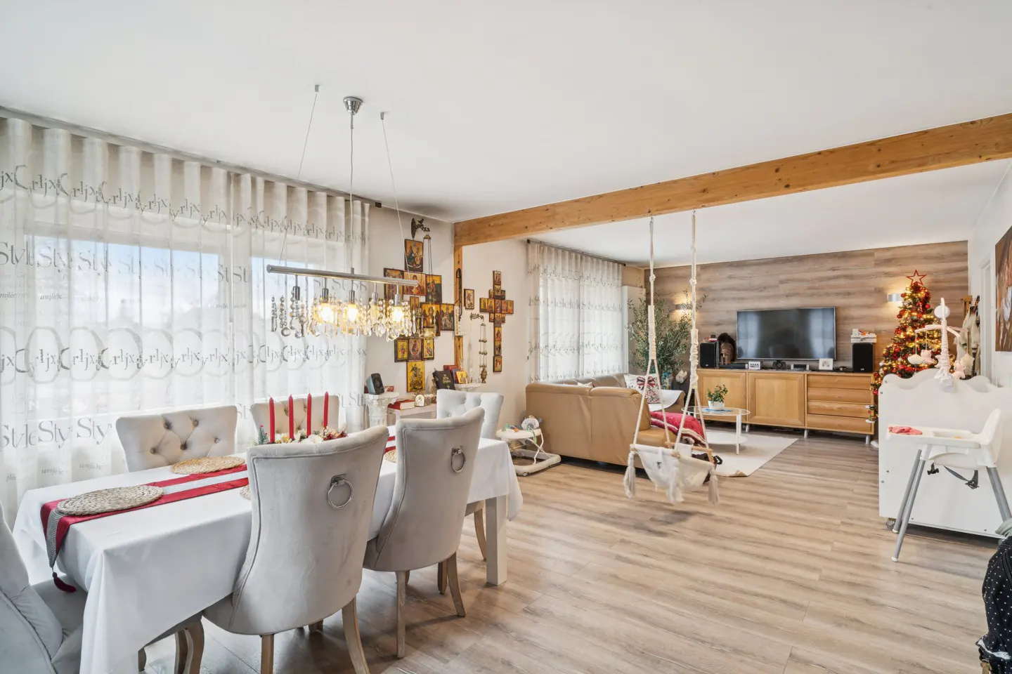 Bright, open-concept living and dining area with light wood floors. Dining table with white tablecloth and gray chairs. Christmas tree in the background.