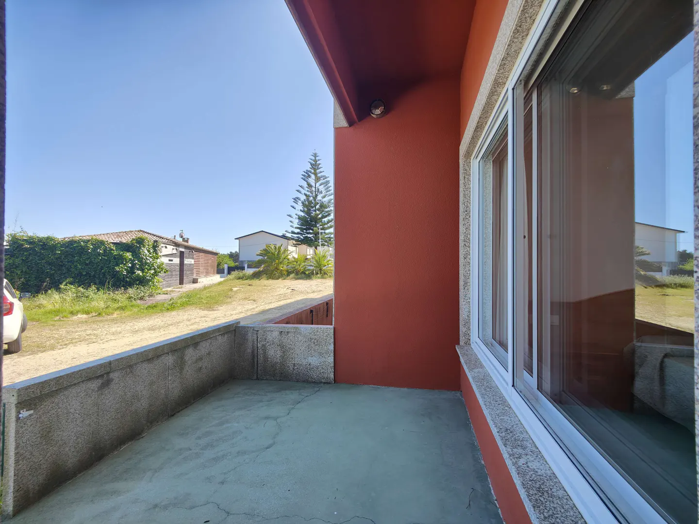 View from a green patio with a low concrete wall, looking out to a yard with houses and a tall pine tree under a blue sky. The building is red with white-framed windows.