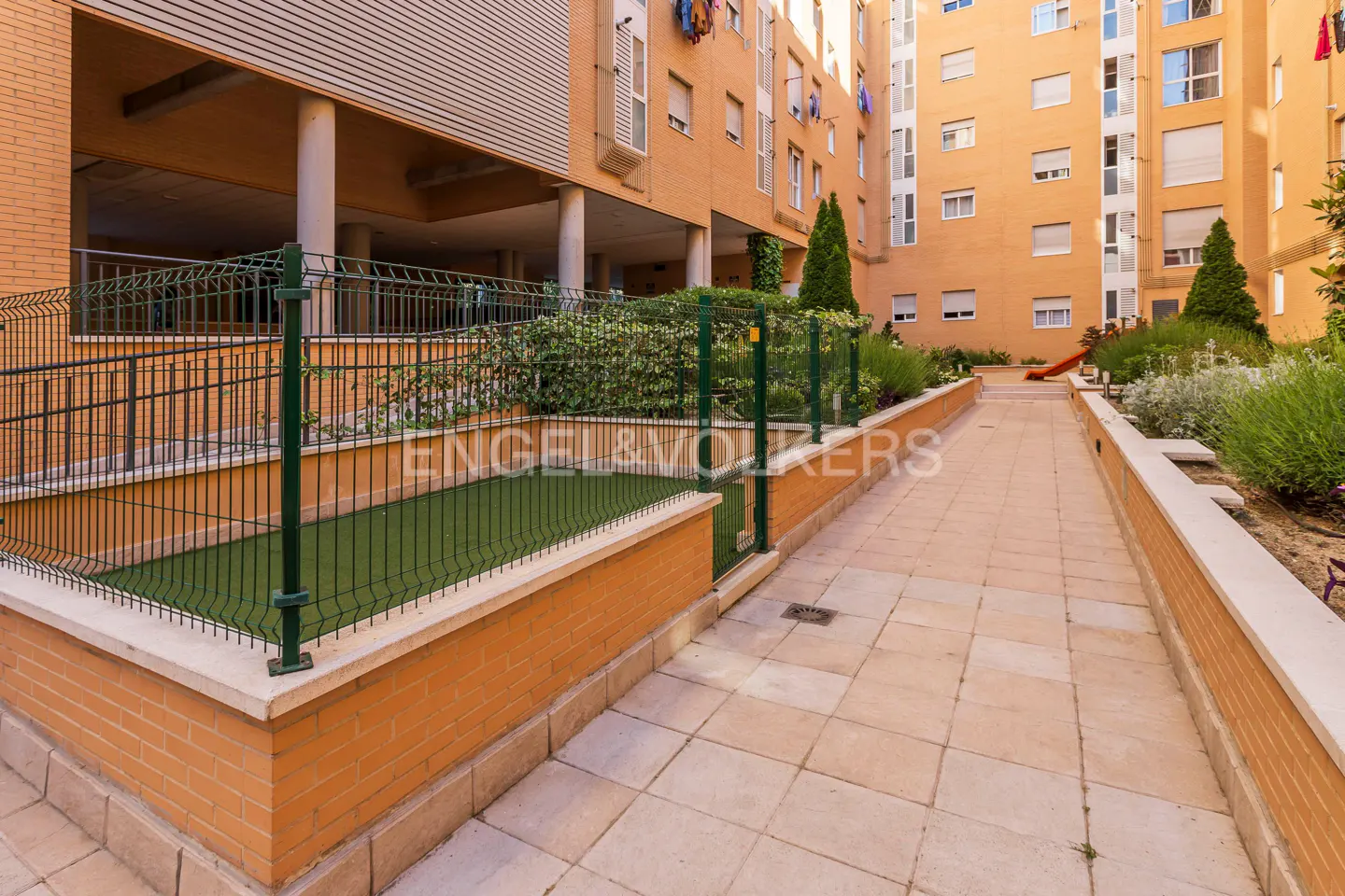 A paved walkway leads past a fenced-in green lawn area next to a tall, orange-brick apartment building with many windows.
