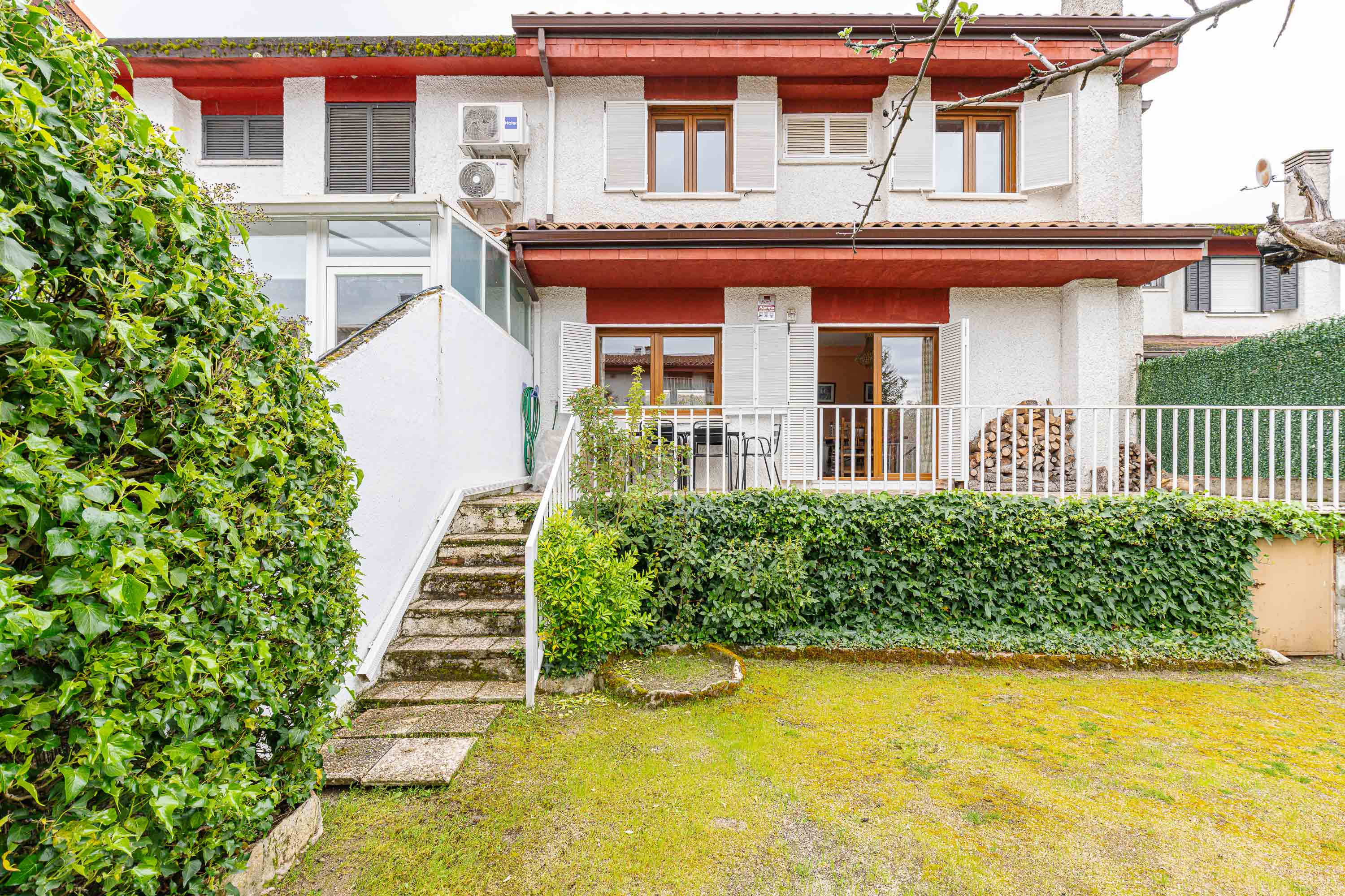Exterior view of a two-story white house with red trim, a small deck, and a green lawn. Stone steps lead to a white wall and a green hose.