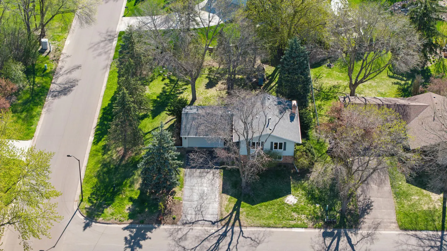 Aerial view of a light blue single-story house with a gray roof, surrounded by green trees and lawn. A concrete driveway leads to the street.