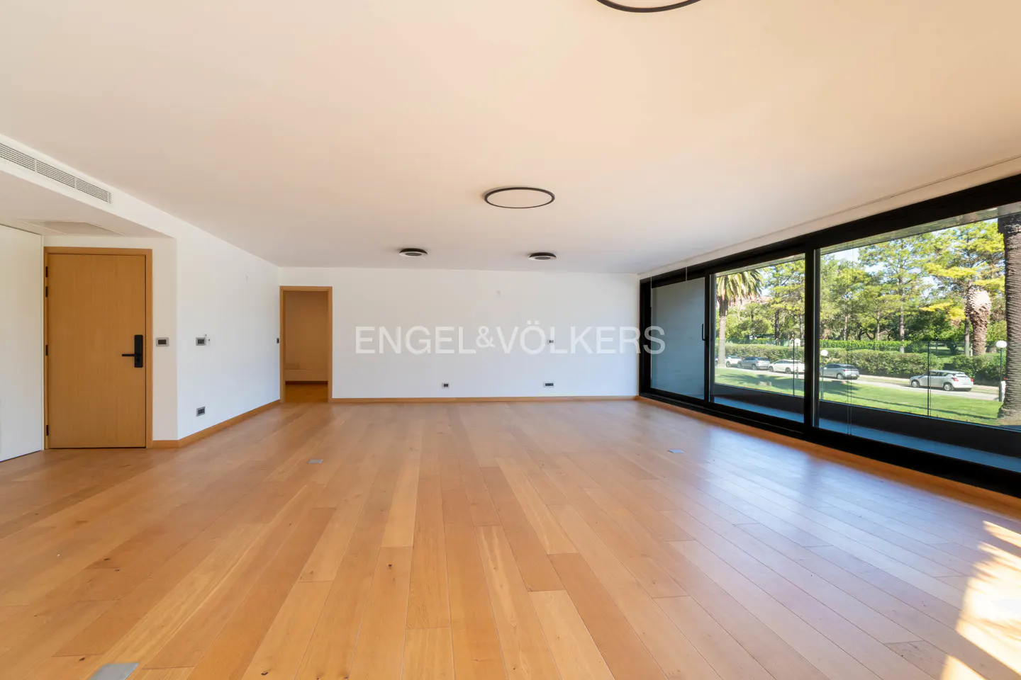 Bright, empty room with wood floors, white walls, and large windows overlooking a green landscape. Door on the left. Engel & Völkers logo.