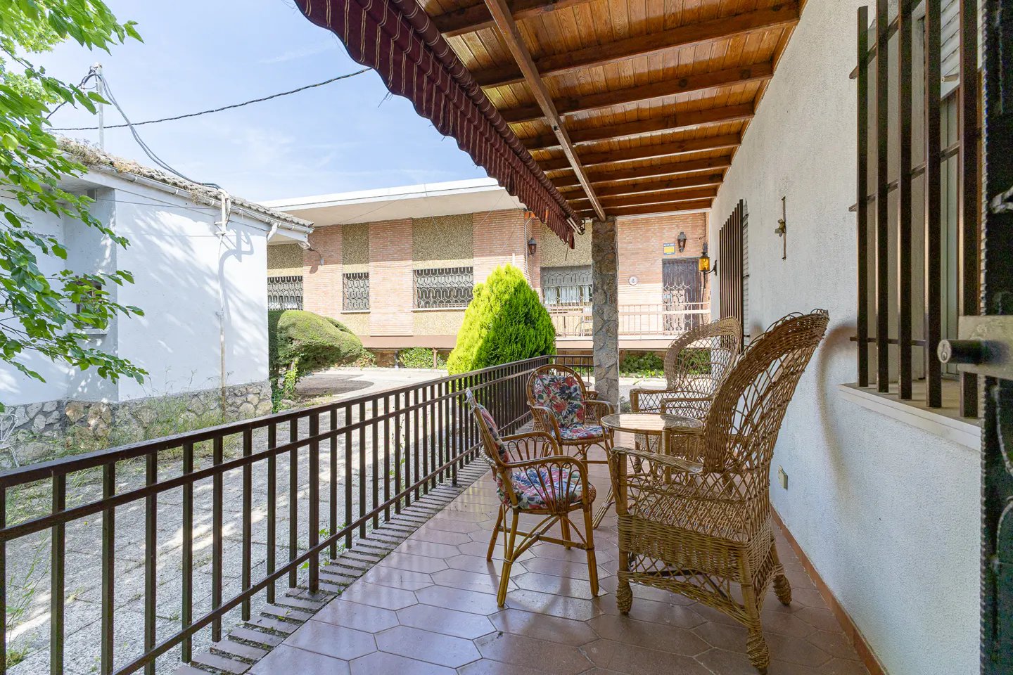 Covered patio with wicker chairs, tiled floor, and metal railing overlooking a sunny courtyard with brick buildings.