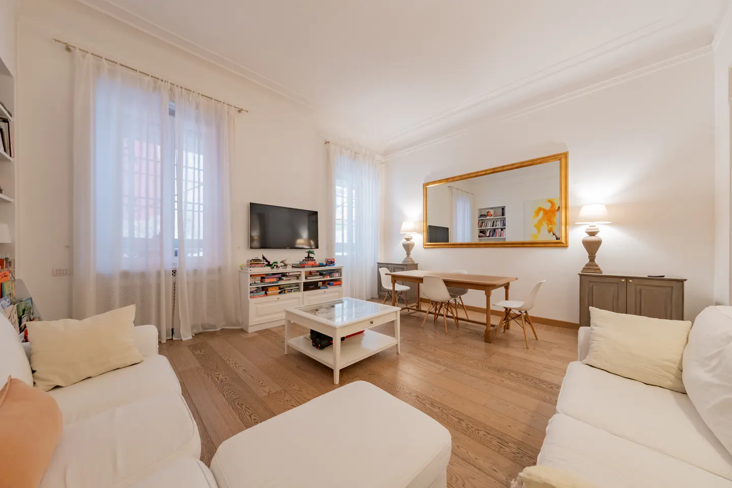 Bright living room with white sofas, wood floors, and a large gold-framed mirror reflecting a dining table and bookshelf.