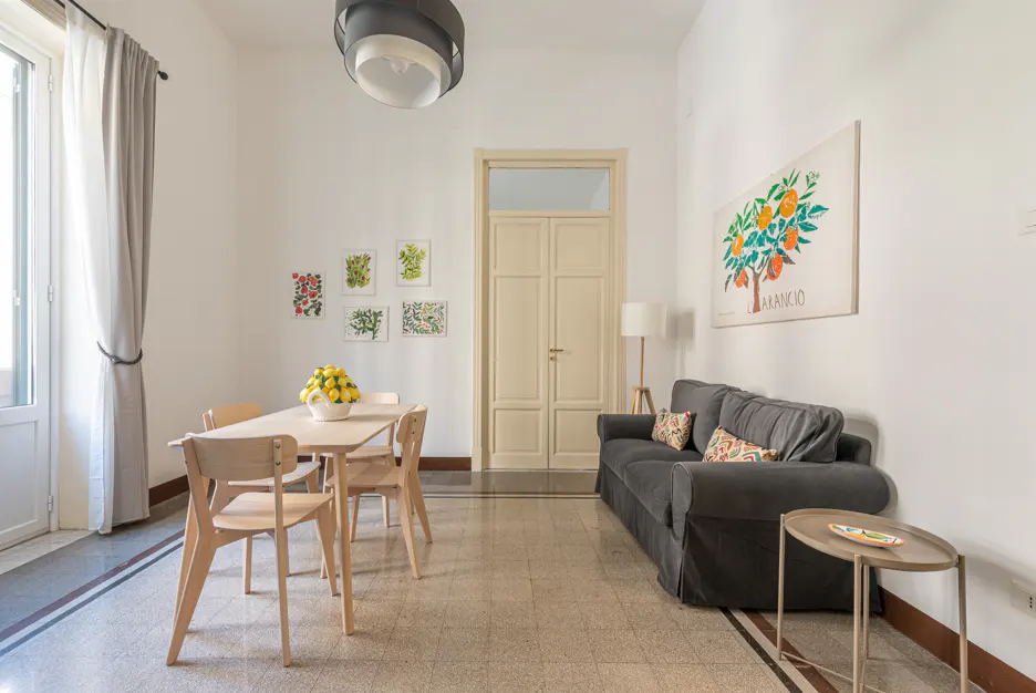 Bright living space with a light wood table, chairs, and a bowl of lemons. A gray sofa sits near a wall with an orange tree print.