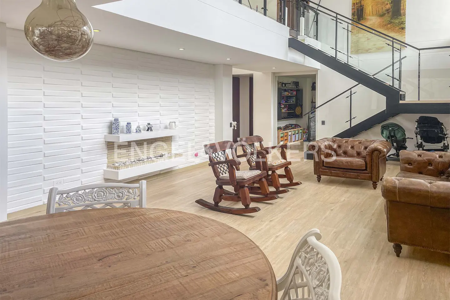 Bright living room with wood floors, a round table, rocking chairs, and brown leather sofas. A modern staircase is visible in the background.