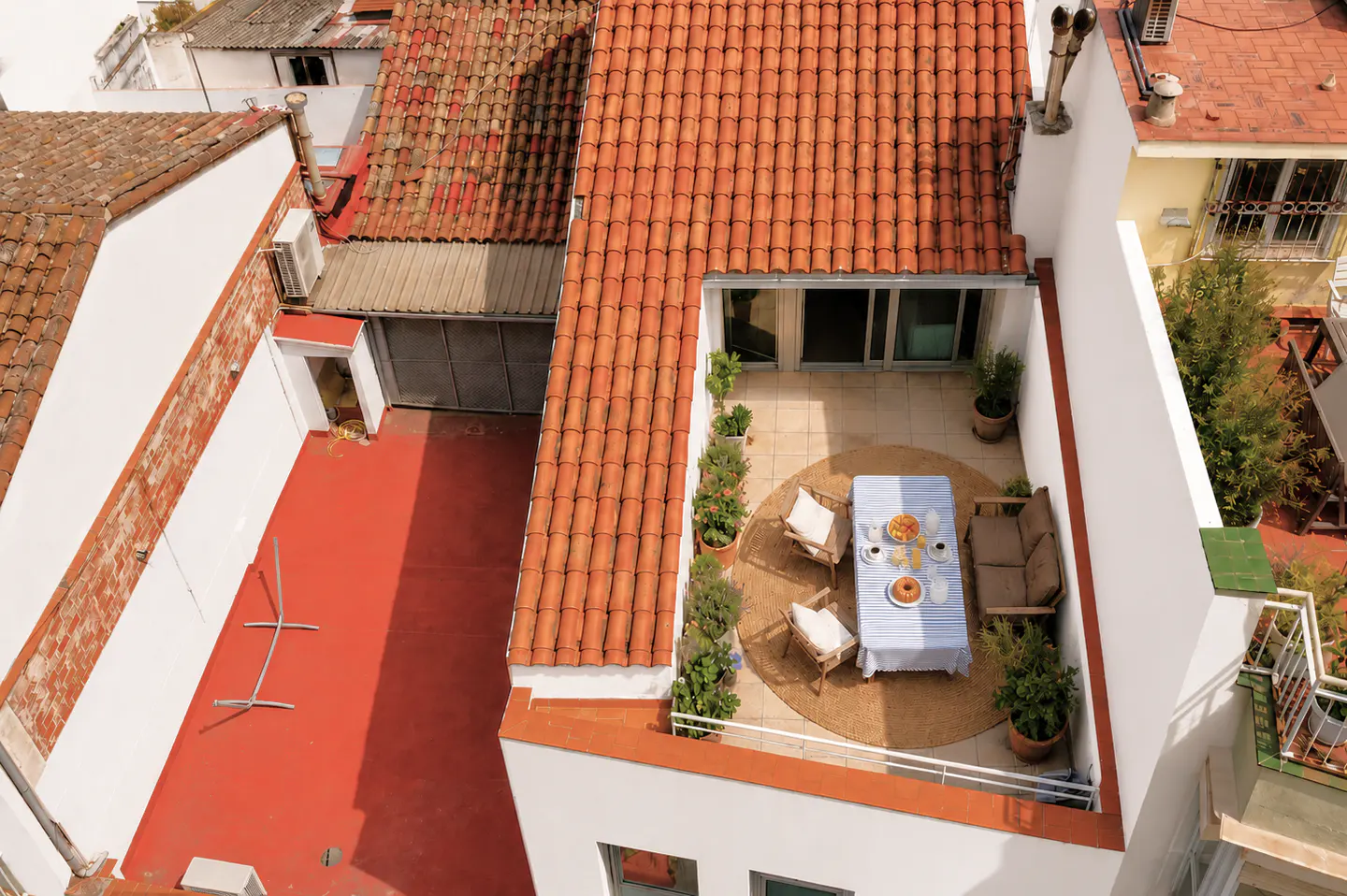 Aerial view of a rooftop terrace with terracotta tiles, a table set for breakfast, and potted plants.