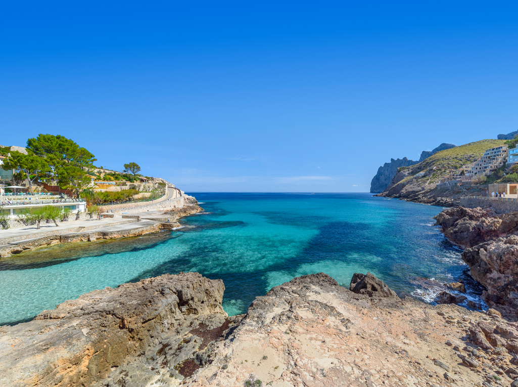 A scenic view of a turquoise Mediterranean cove framed by rocky cliffs, with a coastal path and buildings on the left. [AI-generated text]