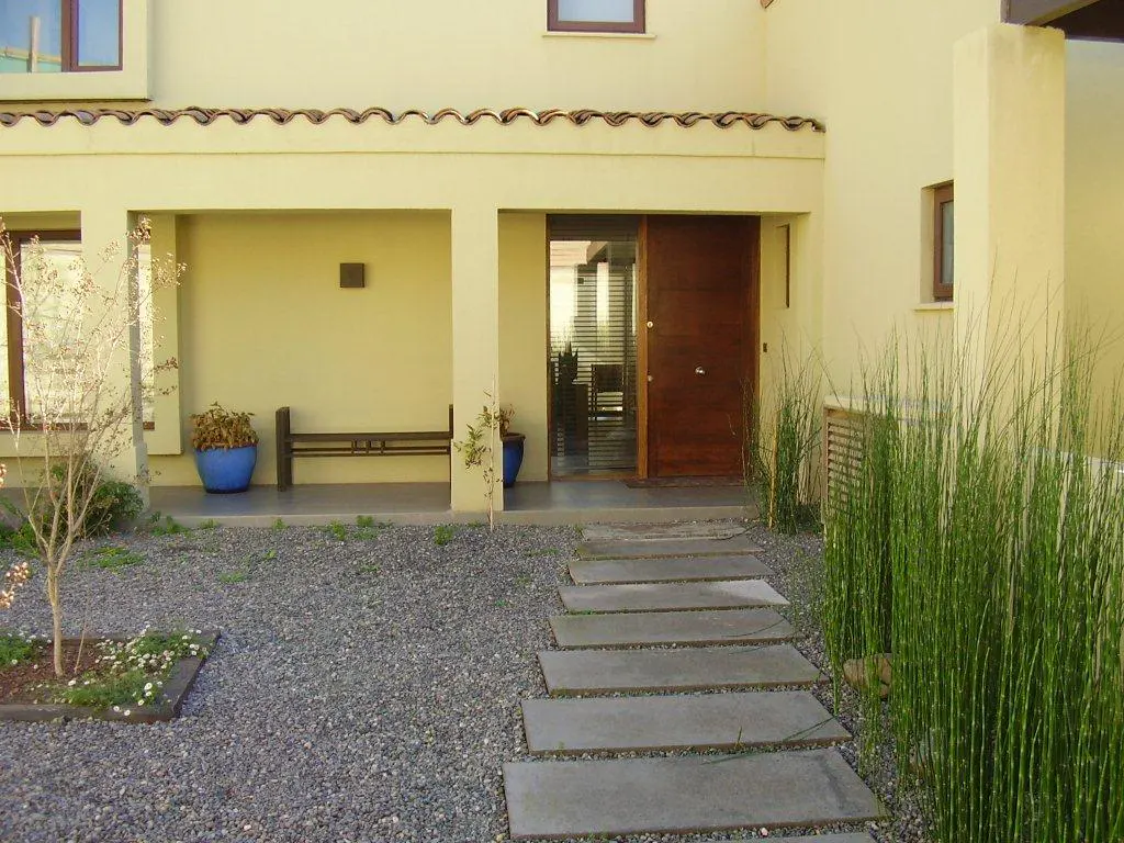 Exterior of a modern home with a stone path leading to a wooden door. A covered porch has a bench and blue planters. Tall grass accents the entrance.