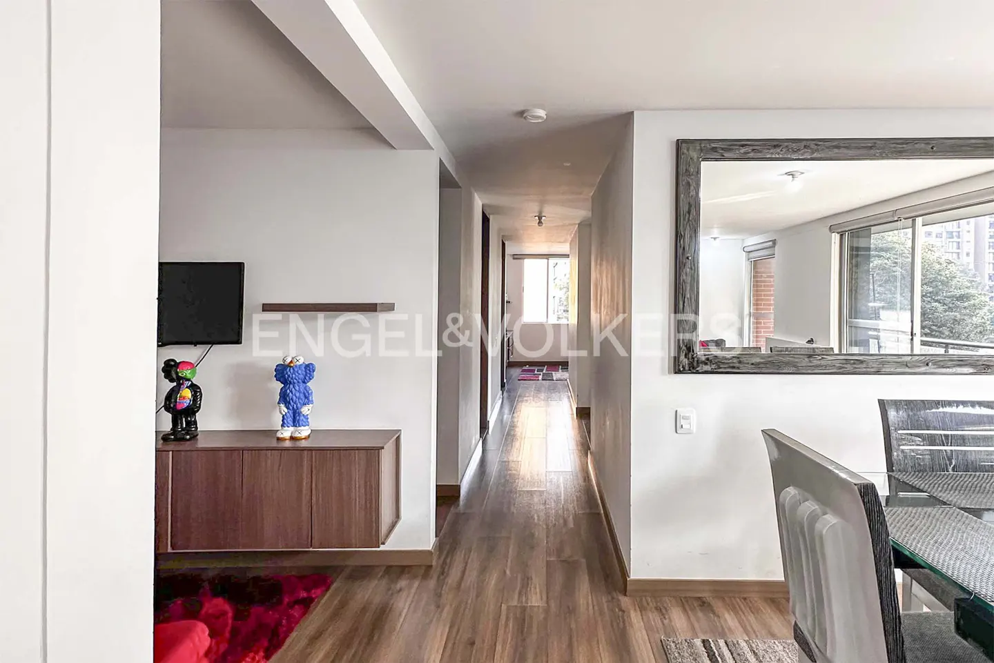 Bright hallway with wood floors, a wooden console table with two art figures, and a large mirror reflecting a dining area.