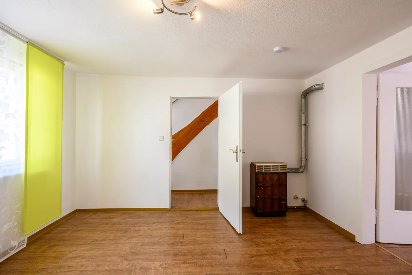 Bright room with wood floors, white walls, and a yellow curtain. An open door reveals a staircase. A vintage cabinet sits near a metal pipe.